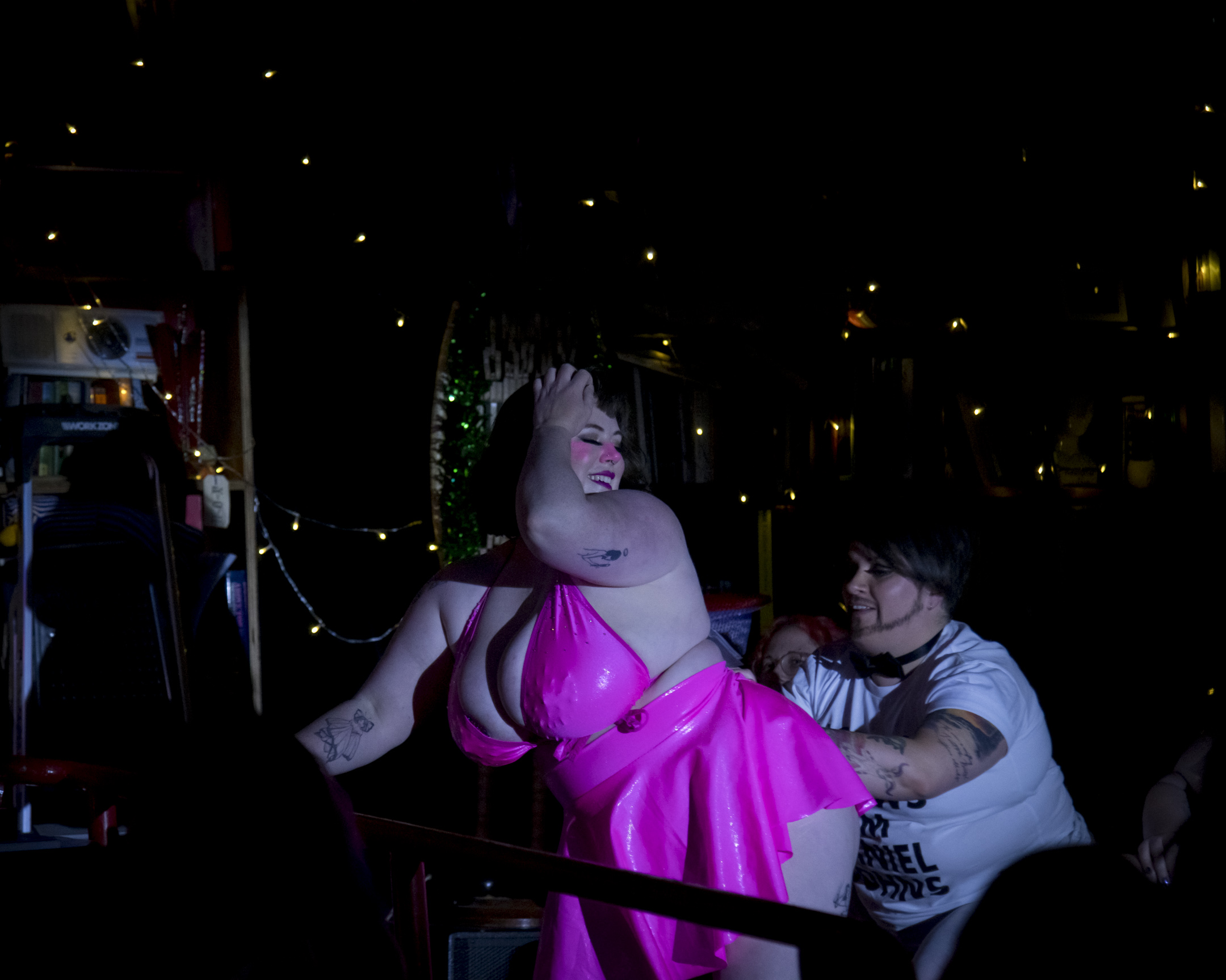 Caitlin Catastrophe - a white woman with short brown hair is performing, wearing a bright pink bikini top and matching mini skirt. An audience member is placing a tipping note into her bikini strap